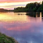 Fisker ved elv under solnedgang i naturen, utendørs aktiviteter, lystfiske, fredelig landskap, miljøvennlig rekreasjon, friluftsliv, sommer, rolig vann, grønne trær, naturopplevelse, avslapning.