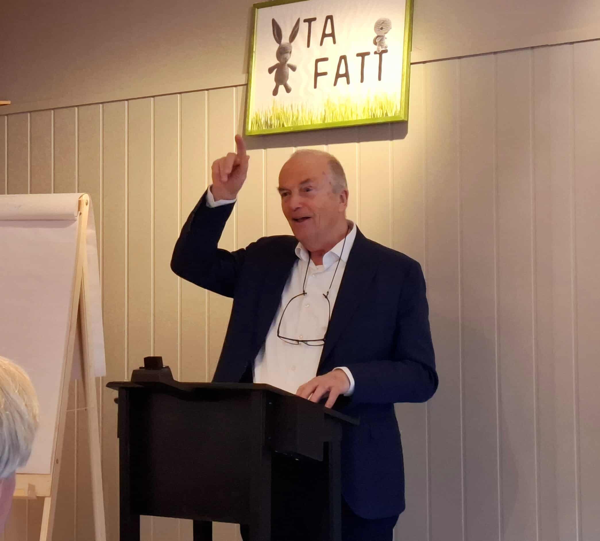 Skarp mann i dress, gestikulerer foran logoet "Ta Fatt" i en konferansesal.