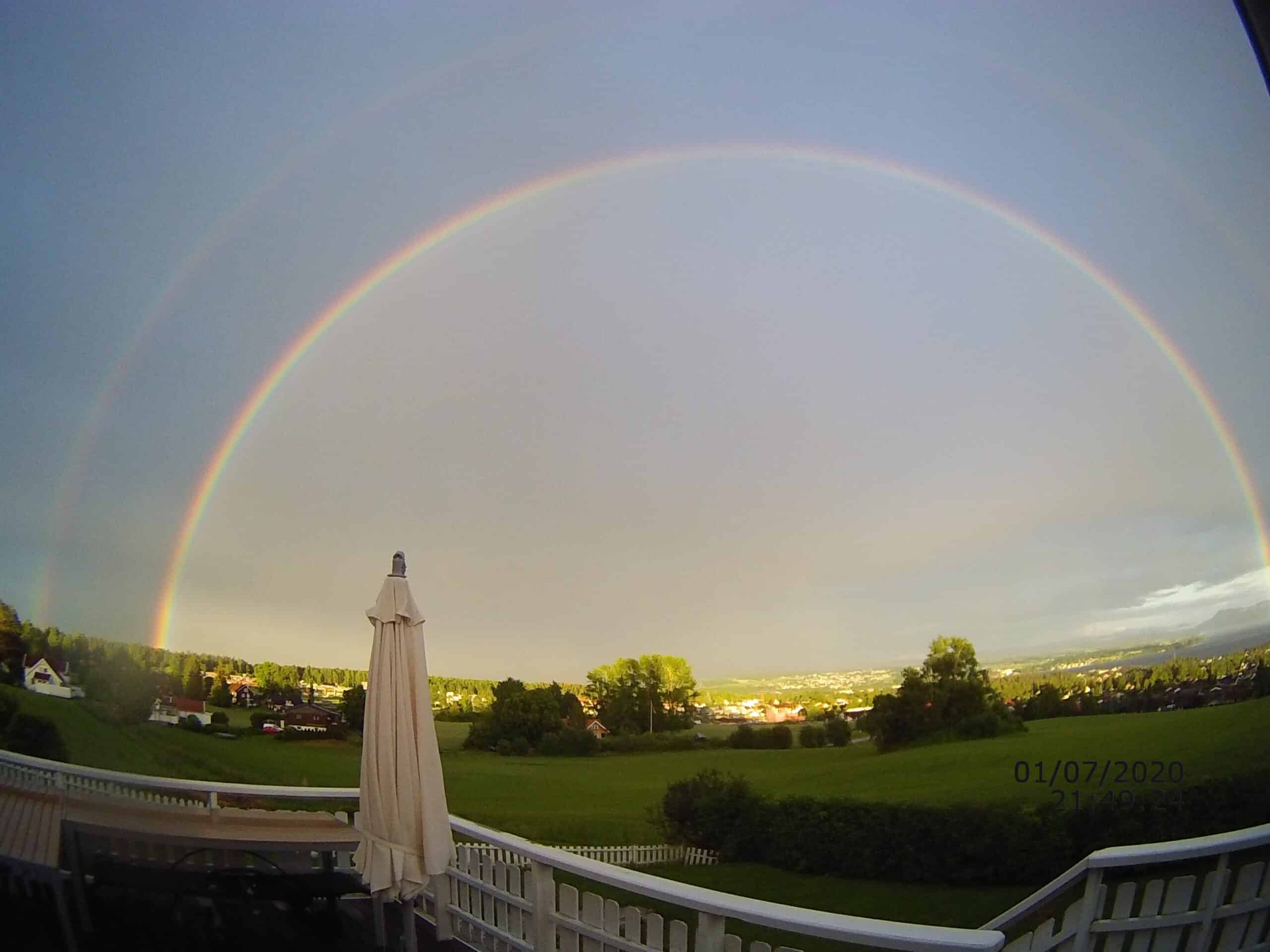 Dobbel regnbue over landskapet sett fra en terrasse med parasoll, med grønne enger og hus i bakgrunnen, under en klar himmel med lette skyer, inspirerende natur og værpalett.
