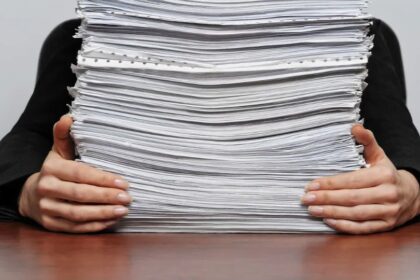 Nesten ferdig med å sortere store mengder papirarbeid for bedriften, teknisk person i svart dress holder en stor bunke med dokumenter. Fokus på administrasjon, arkiv, og kontorarbeid.