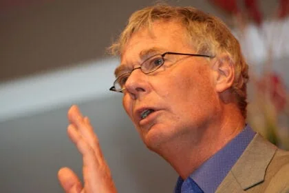 Kraftig mann i briller som holder en hånd opp under presentasjon eller tale. Fokus på ansikt og overkropp, bruk av blazer og skjorte, bakgrunn med fargerik dekorasjon.