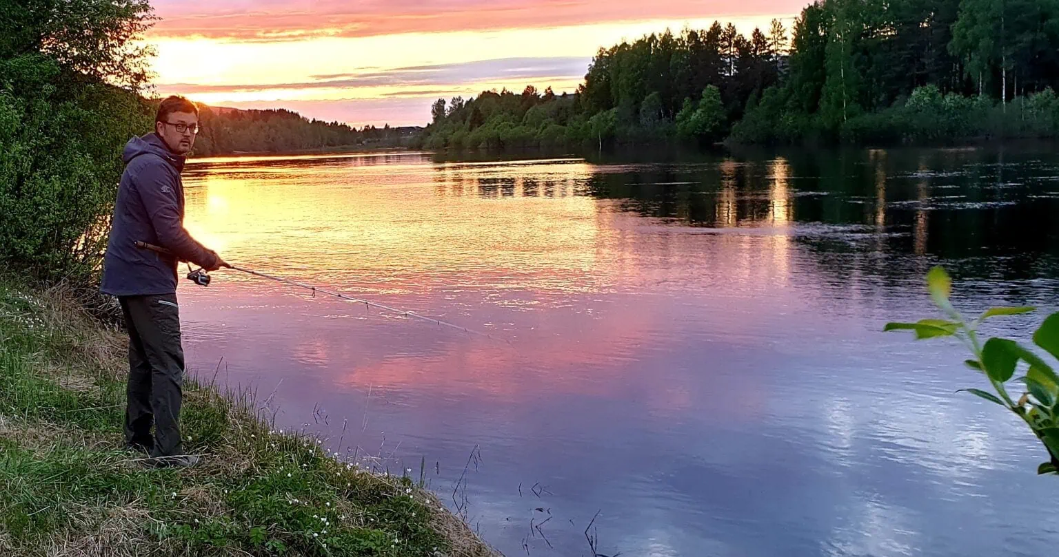 Fisker ved elv under solnedgang i naturen, utendørs aktiviteter, lystfiske, fredelig landskap, miljøvennlig rekreasjon, friluftsliv, sommer, rolig vann, grønne trær, naturopplevelse, avslapning.