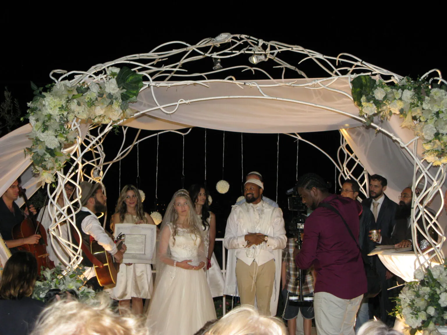 Festlig utendørs bryllupsseremoni under en lyssatt blomsterdekorert pergola, med brud, forlover og musikere, skaper en romantisk atmosfære for den spesielle dagen.