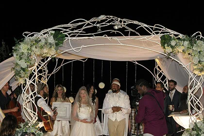 Festlig utendørs bryllupsseremoni under en lyssatt blomsterdekorert pergola, med brud, forlover og musikere, skaper en romantisk atmosfære for den spesielle dagen.