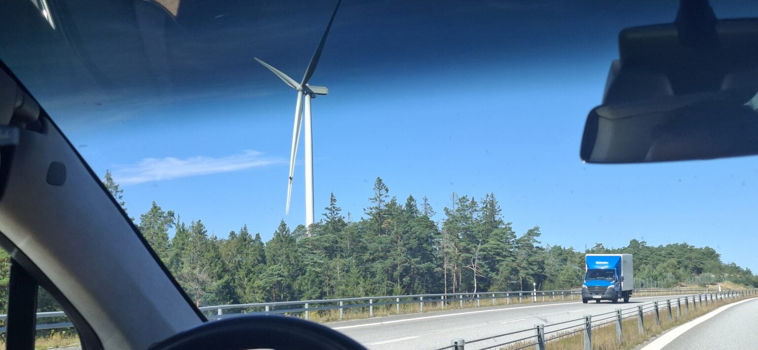 Vindmølle på landskap med blå himmel sett fra bilvindu, motorvei og trær i bakgrunnen.