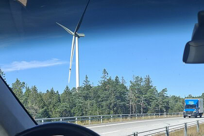 Vindmølle på landskap med blå himmel sett fra bilvindu, motorvei og trær i bakgrunnen.