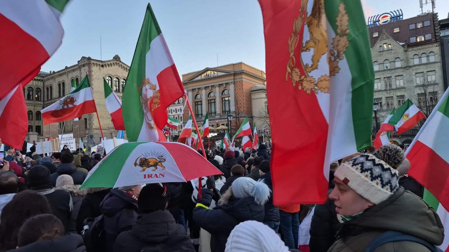 Fargerike norske og iranske flagg i en folkemengde under en utendørs demonstrasjon eller feiring, med historiske bygninger i bakgrunnen.