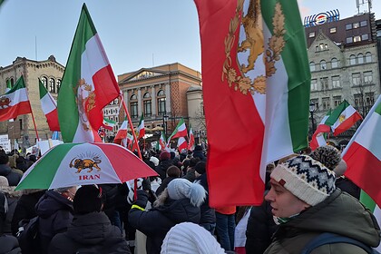 Fargerike norske og iranske flagg i en folkemengde under en utendørs demonstrasjon eller feiring, med historiske bygninger i bakgrunnen.